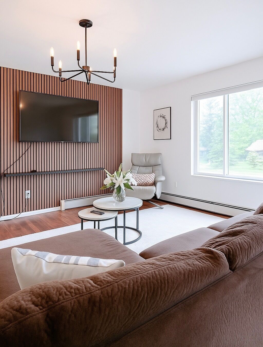 A modern living room with a large flat-screen TV mounted on a wooden wall, a comfortable sofa, a round coffee table with a vase of flowers, and a large window providing a view of the outdoors.