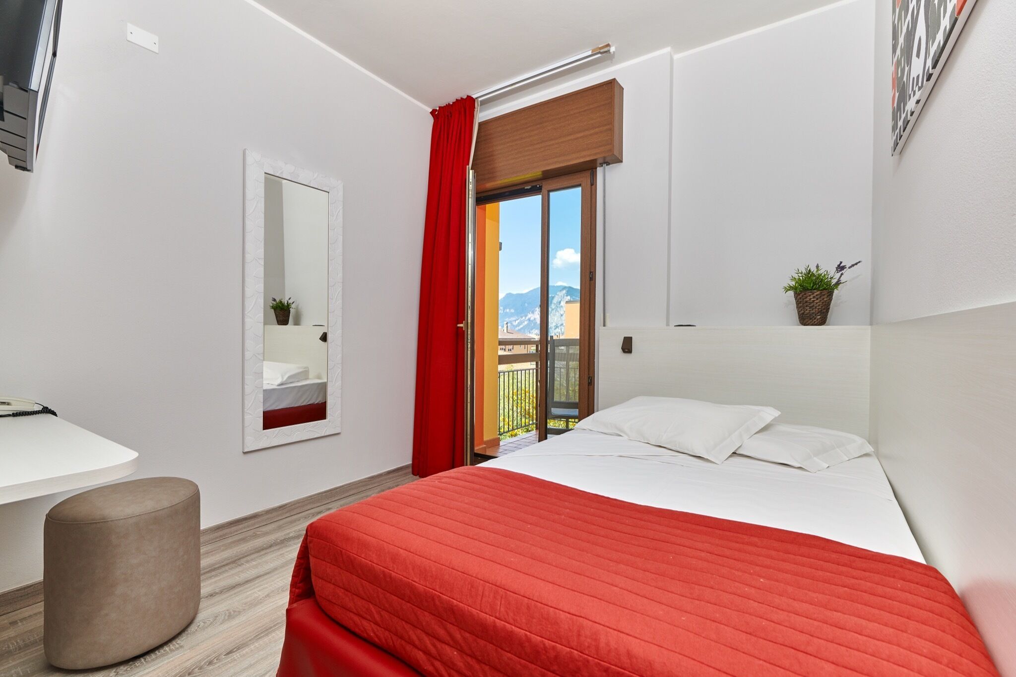 Une chambre d’hôtel moderne équipée d’un lit, d’un bureau, d’un miroir et d’un balcon avec un rideau rouge.