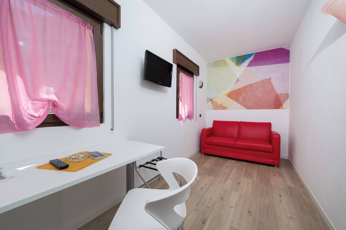 Quarto moderno com escrivaninha branca, sofá vermelho e arte abstrata colorida na parede.