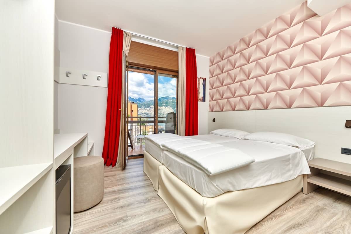 Quarto de hotel moderno com cama, escrivaninha, cadeira e varanda com cortinas vermelhas.