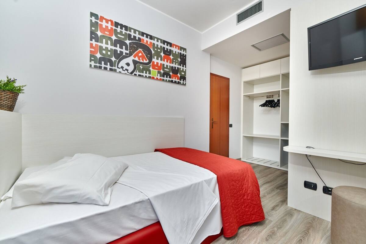 Quarto de hotel moderno com cama, parede de destaque vermelha, televisão presa à parede e guarda-roupa com espaço para pendurar roupas.