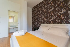 A bedroom with a bed, a bedside table, a nightstand, a large mirror, and a bathroom visible through an open door.