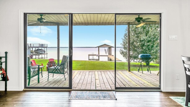 Une pièce dotée d’une grande porte-fenêtre coulissante donnant sur une terrasse extérieure avec un barbecue, des chaises et une vue sur une étendue d’eau.