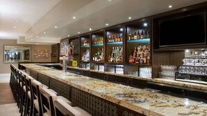 A bar area with a long counter, multiple shelves of bottles, and a television screen mounted on the wall.
