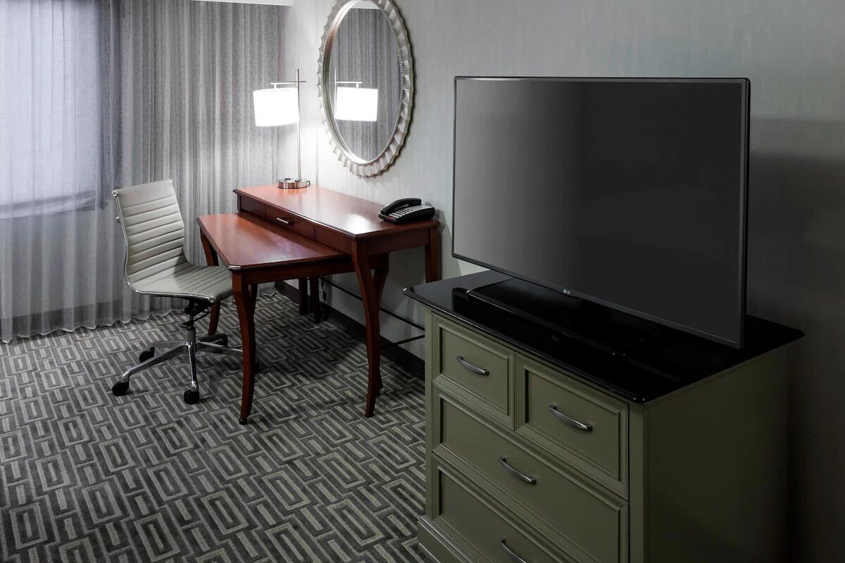 A hotel room with a desk, chair, television, and a minibar.