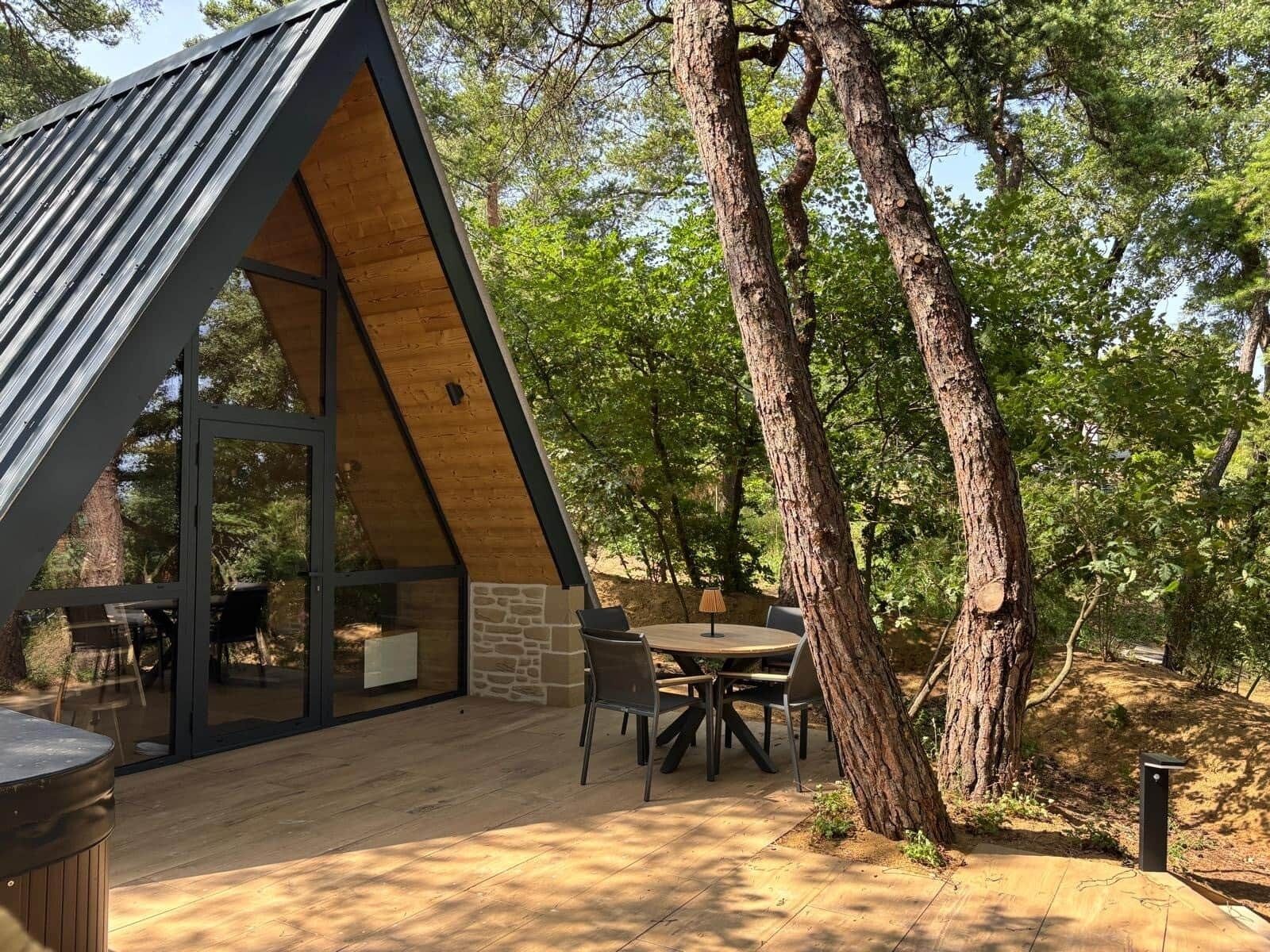 A modern, A-frame wooden structure with a glass door, a stone wall, and a patio with a table and chairs.