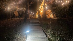 A wooden pathway leading to a modern house with string lights and a large woven chair. - CollinéA Chalet "CanopéA" 28 m² Private SPA (Bathernay)