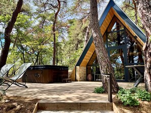 Une cabane avec un toit en pente, entourée d’arbres, et un espace patio comprenant une chaise et un jacuzzi.