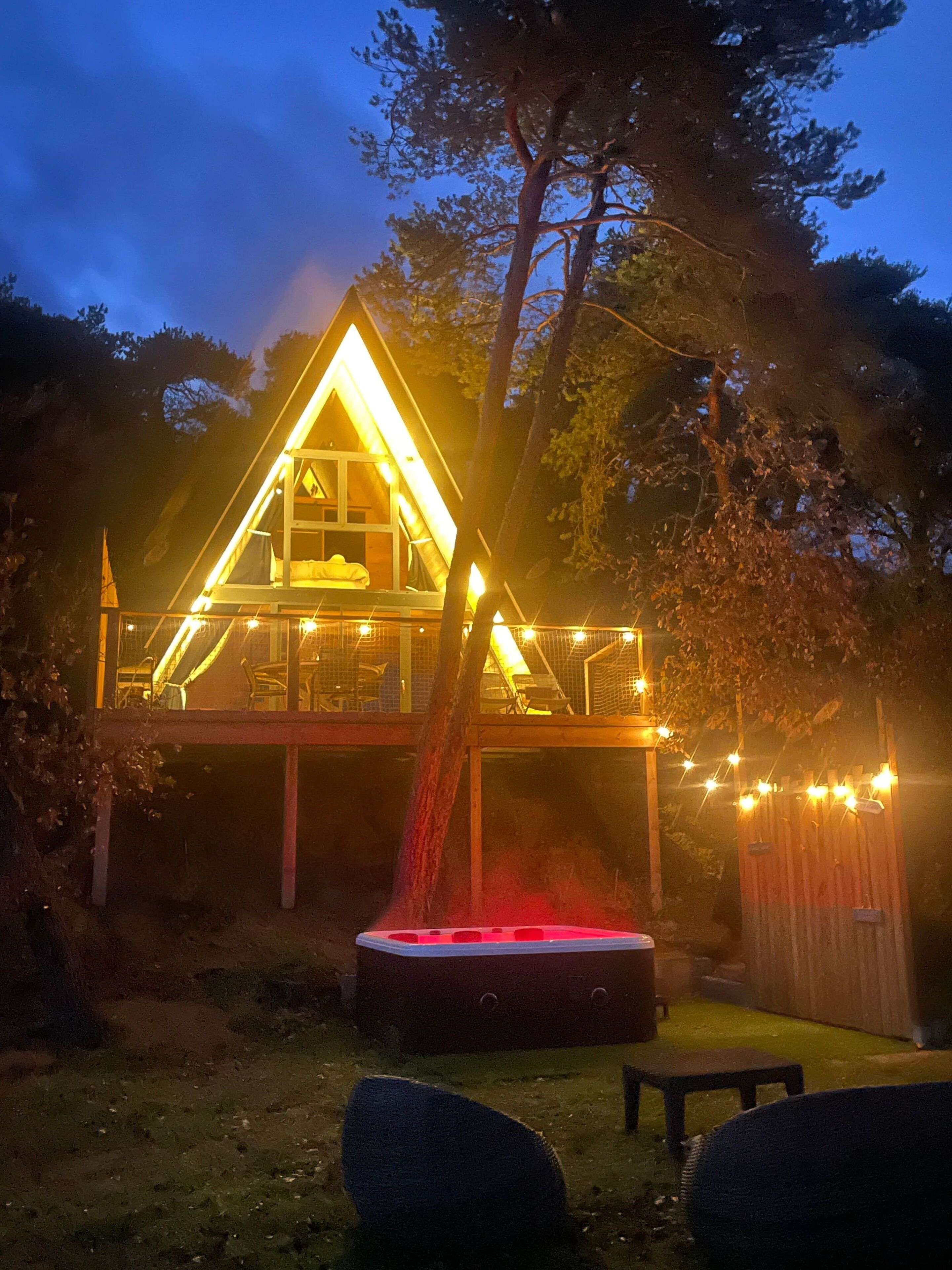 A triangular wooden structure with a glass upper level, surrounded by trees and a string of lights.
