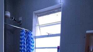 Une petite salle de bain avec des toilettes blanches, un espace douche doté d’un rideau de douche à rayures bleues et blanches, une fenêtre au-dessus des toilettes et un lavabo avec un distributeur de savon.