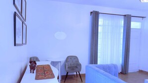 Un salon avec un canapé bleu, une table basse blanche et un fauteuil gris. Il y a une fenêtre avec des rideaux et une lampe fixée au mur.
