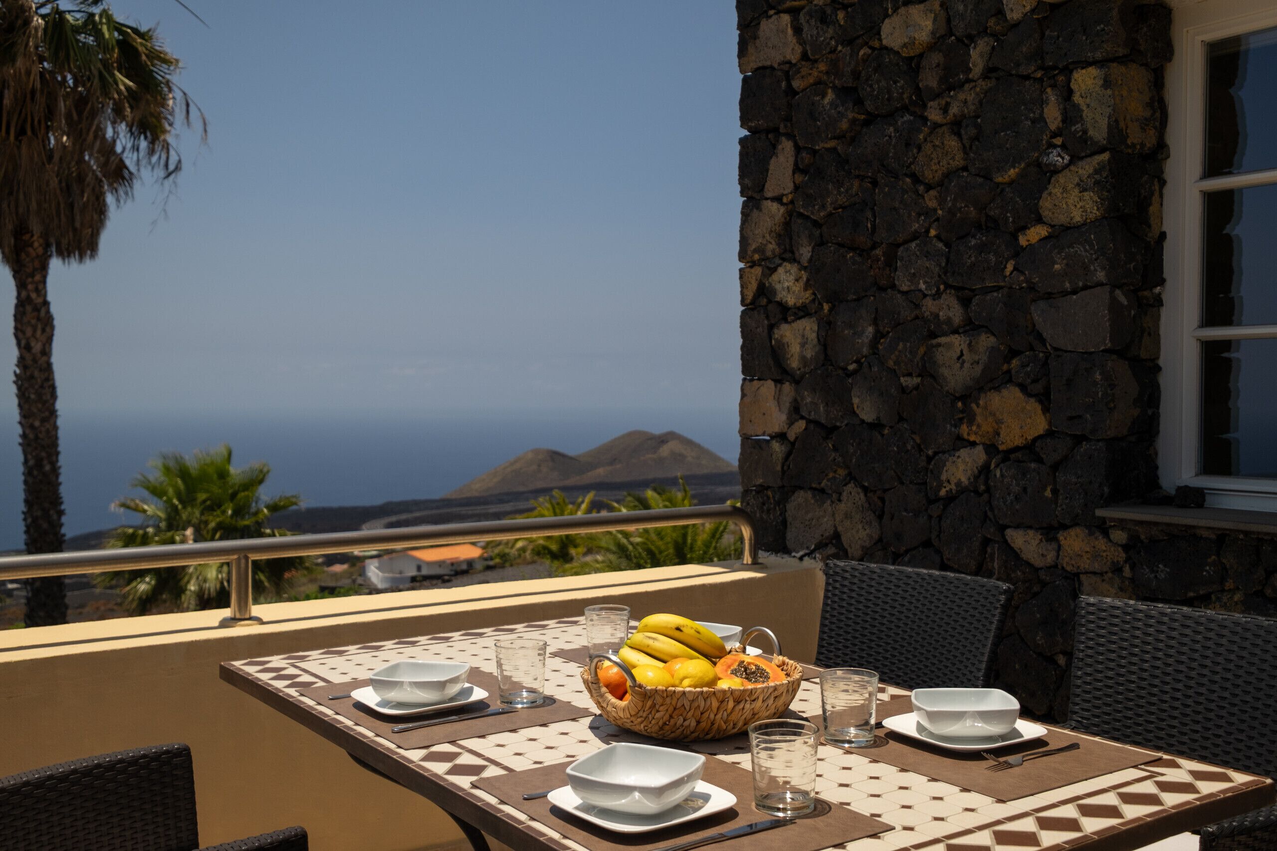 A table with a fruit basket, bowls, and glasses on an outdoor balcony with a stone wall and a view of the ocean.