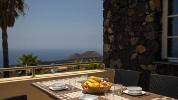 A table with a fruit basket, bowls, and glasses on an outdoor balcony with a stone wall and a view of the ocean.