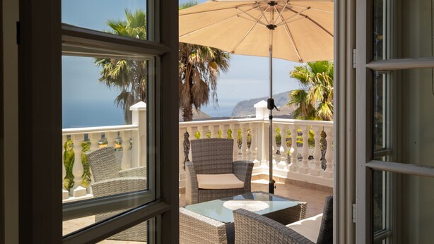 Ein Balkon mit Korbmöbeln, einem Glastisch und einem beige-farbenen Sonnenschirm.