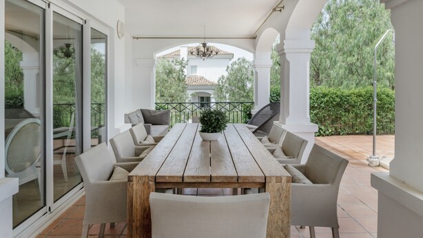 Een overdekt terras met een houten tafel en zes stoelen, een glazen deur naar buiten en een uitzicht op een huis op de achtergrond.