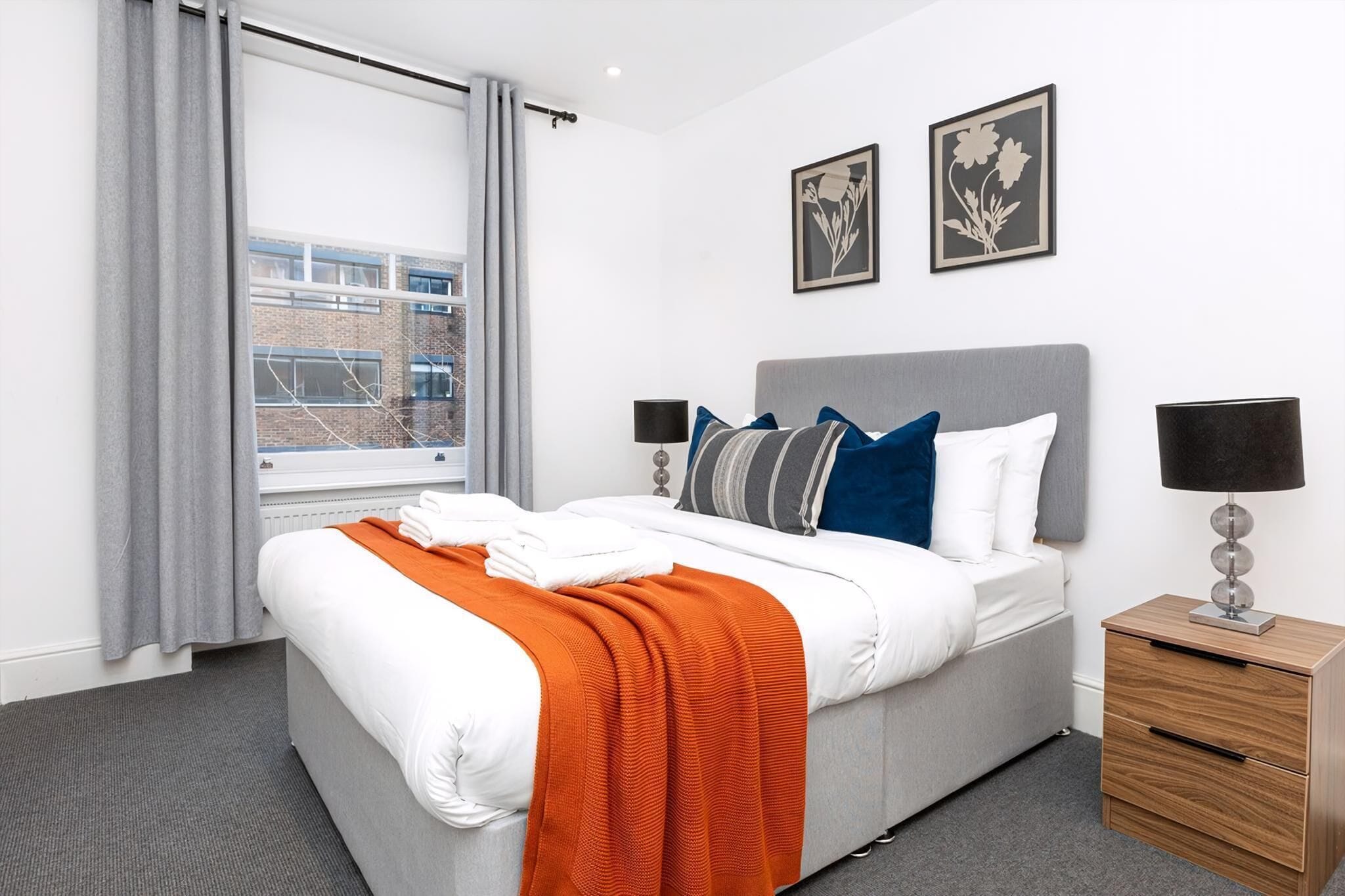 Una habitación de hotel moderna con una cama grande, cortinas grises, vistas a un edificio y dos cuadros enmarcados en la pared.