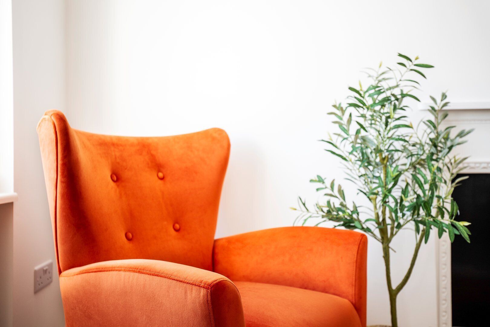 Un fauteuil orange, avec un dossier haut et des motifs capitonnés, placé à côté d’un mur blanc et d’une plante verte.