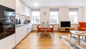 A modern kitchen with white cabinets, a dining area with a wooden table and orange chairs, a television, and a clock on the wall.