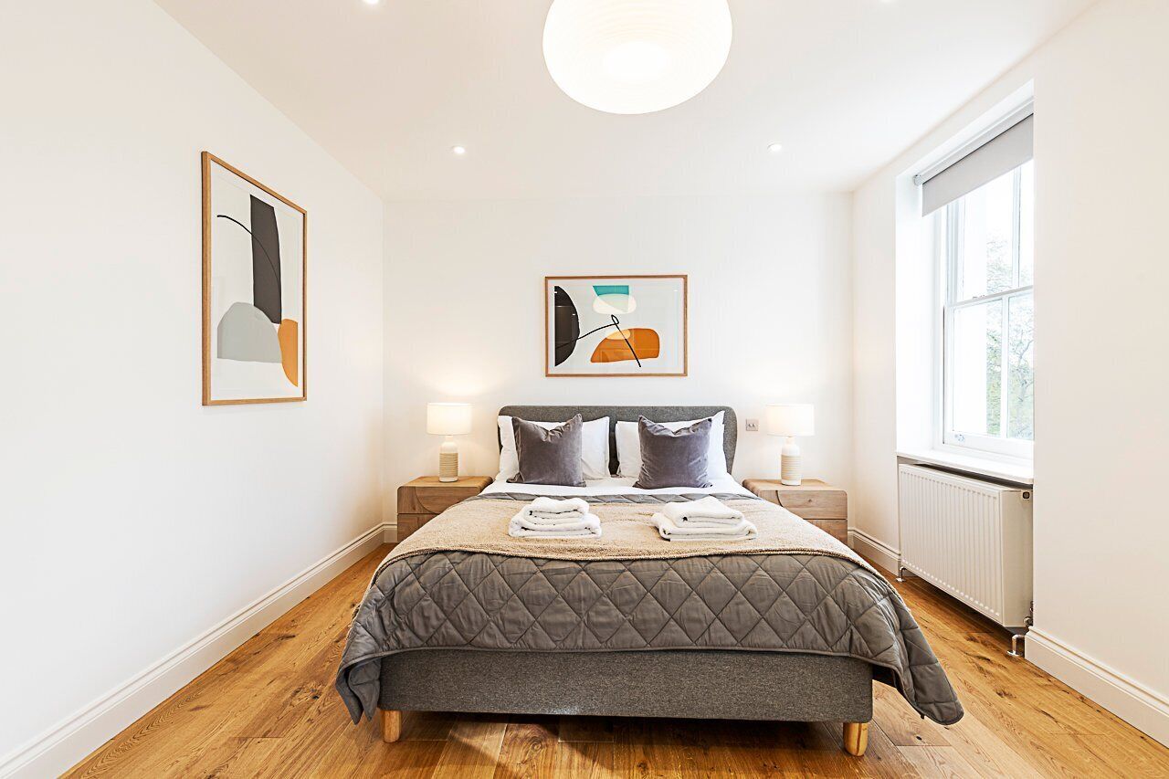 A bedroom with a bed, two bedside tables with lamps, a window, and two framed artworks on the walls.