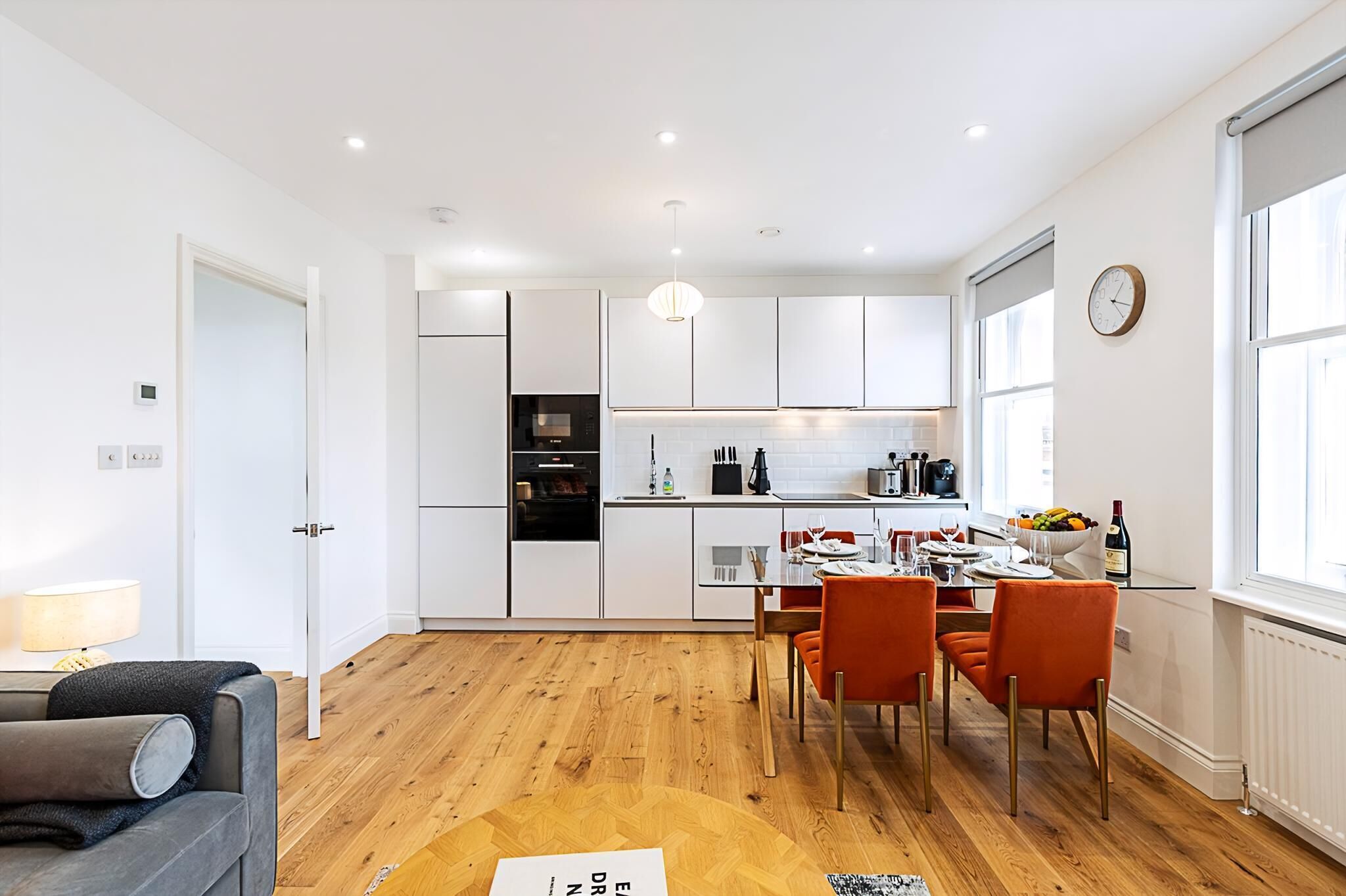 Una cocina moderna con armarios blancos, una zona de comedor con sillas naranjas y suelo de madera.
