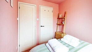 A bedroom with pink walls, two white doors, a wooden dresser, and a bed with a striped bedspread. - Bright 2-bed Home Near Putney Riverside (London)