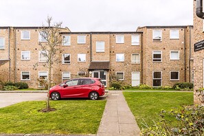 A red car parked in front of a brick apartment building with multiple windows and a small tree in the front yard.