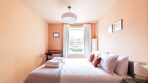 Un dormitorio con cama, escritorio, silla, lámpara y ventana con cortinas.