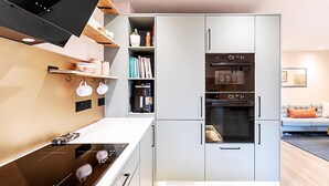 Una cocina moderna con horno empotrado, microondas y cafetera. Se observan estantes con libros y utensilios de cocina.