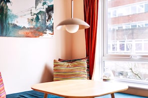 A room with a wooden table, colorful striped pillows, a white pendant light, and a window with a view of buildings and trees.