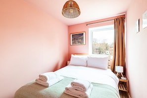 A hotel room with a bed, a window with curtains, a wall-mounted picture, and a bedside table with a lamp.
