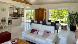 A modern living room with a white sofa, a wooden dining table, and a kitchen area with a sliding door to the outdoors. - House Sainte-Marie de Ré (Sainte-Marie-de-Ré)