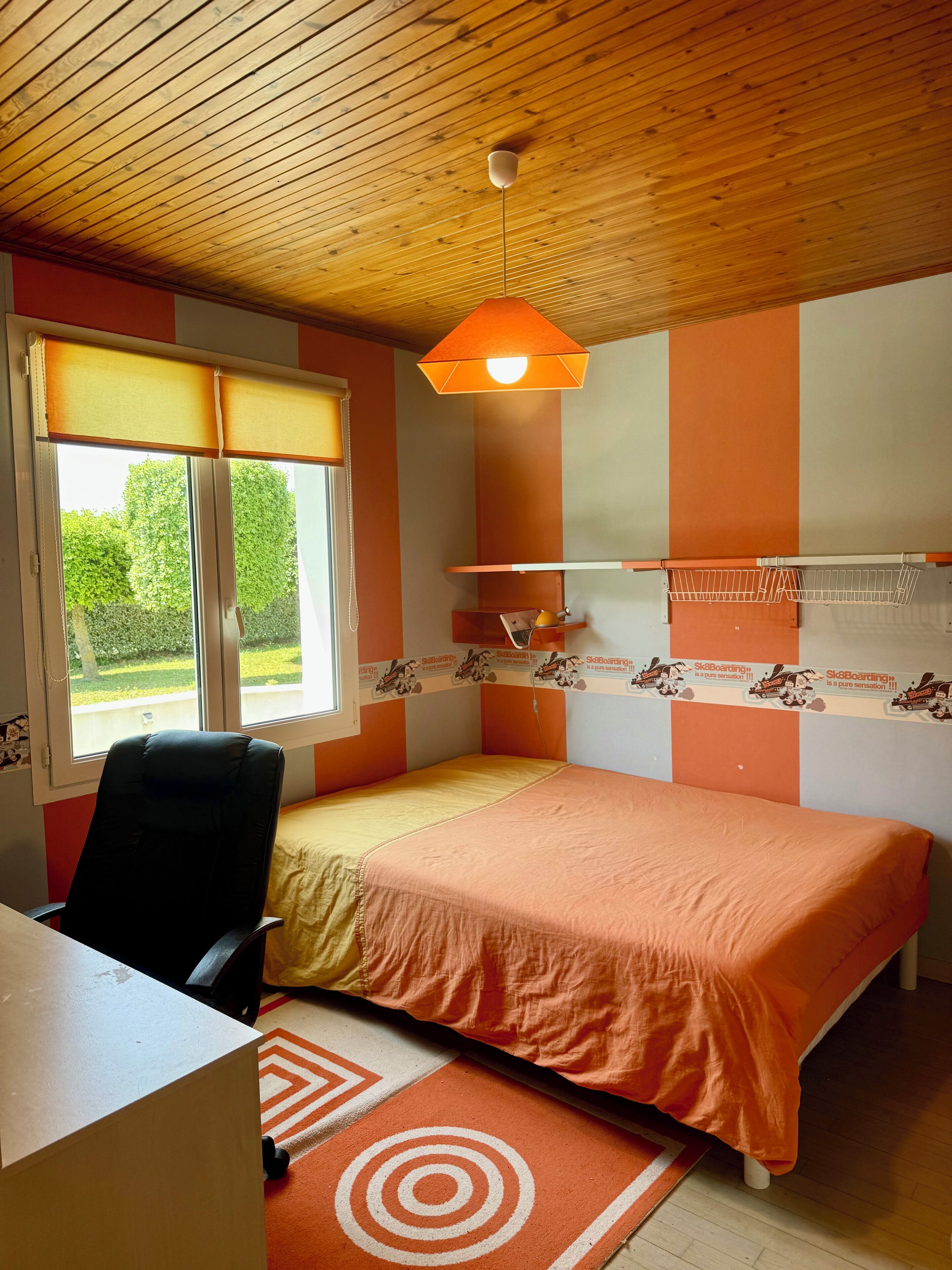 Une chambre avec un lit, un bureau et une chaise. Il y a une fenêtre avec des stores, un ventilateur de plafond et une étagère fixée au mur avec une lampe.