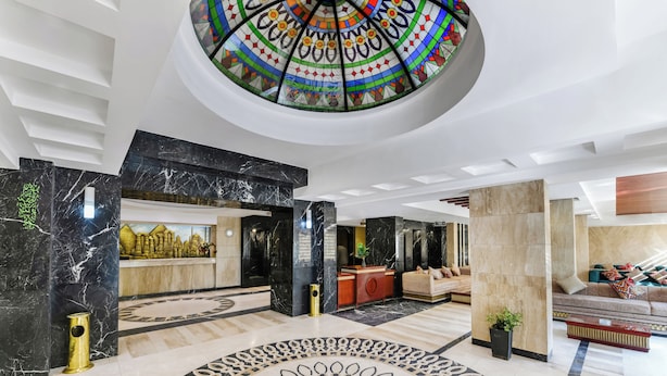 Un hall d’entrée spacieux doté d’un sol circulaire orné de motifs, un espace de réception agrémenté d’une fresque représentant un paysage urbain, et un coin salon avec des canapés et une table basse.