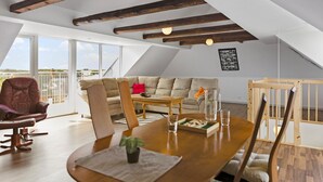 Ein modernes Loft mit freiliegenden Holzbalken, einem großen Fenster, einer Couch, einem Esstisch und Blick ins Freie.