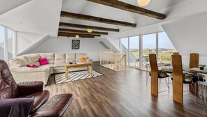 Ein geräumiger Loft mit hohen Decken, freiliegenden Holzbalken, einem großen Fenster, einer gemütlichen Couch, einem Essbereich und Blick auf die Landschaft.