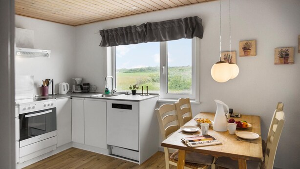 Eine moderne Küche mit weißen Schränken, einem Herd und einem Geschirrspüler. Ein Esstisch mit Stühlen und ein Fenster mit Blick ins Grüne.