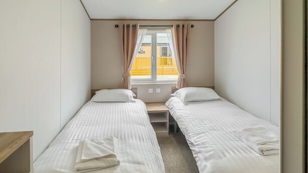 Une chambre avec deux lits simples, une fenêtre avec des rideaux, un banc en bois et une vue sur un immeuble jaune à l’extérieur.