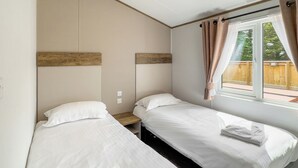 Une chambre avec deux lits, une fenêtre avec des rideaux, une tête de lit en bois et une petite table de chevet en bois.