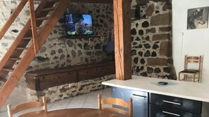 Een landelijke keuken met een stenen muur, houten balken, een ronde houten tafel met stoelen, een aan de muur bevestigde televisie en een houten stoel.