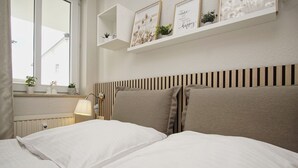 Un lit bien fait, avec une literie blanche, une lampe de chevet et une fenêtre donnant sur des immeubles.