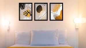 Een hotelkamer met een bed, witte kussens, een gele bedspreide met een patroon en drie ingelijste kunstwerken aan de muur.