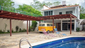 Une piscine avec une terrasse en bois, un fourgon et une maison avec un toit en tuiles.