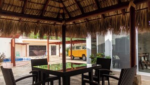 Un espace extérieur abrité, doté d’un toit de chaume, avec une piscine et un véhicule jaune en arrière-plan.