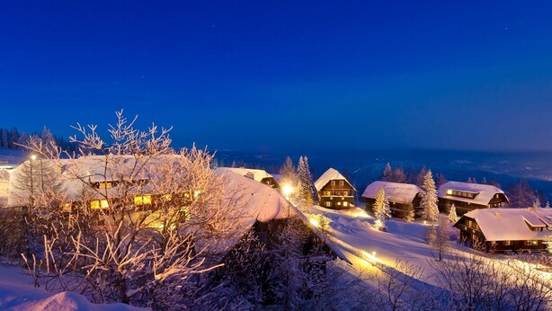 A snow-covered landscape with several wooden cabins illuminated by warm lights, set against a twilight sky. - Apartment by Lake Ossiach with Mountain Views (Sattendorf)