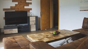 A modern living room with a flat-screen TV, wooden wall decor, a brown leather sofa, and a coffee table with a bowl of fruit. - Apartment by Lake Ossiach with Mountain Views (Sattendorf)