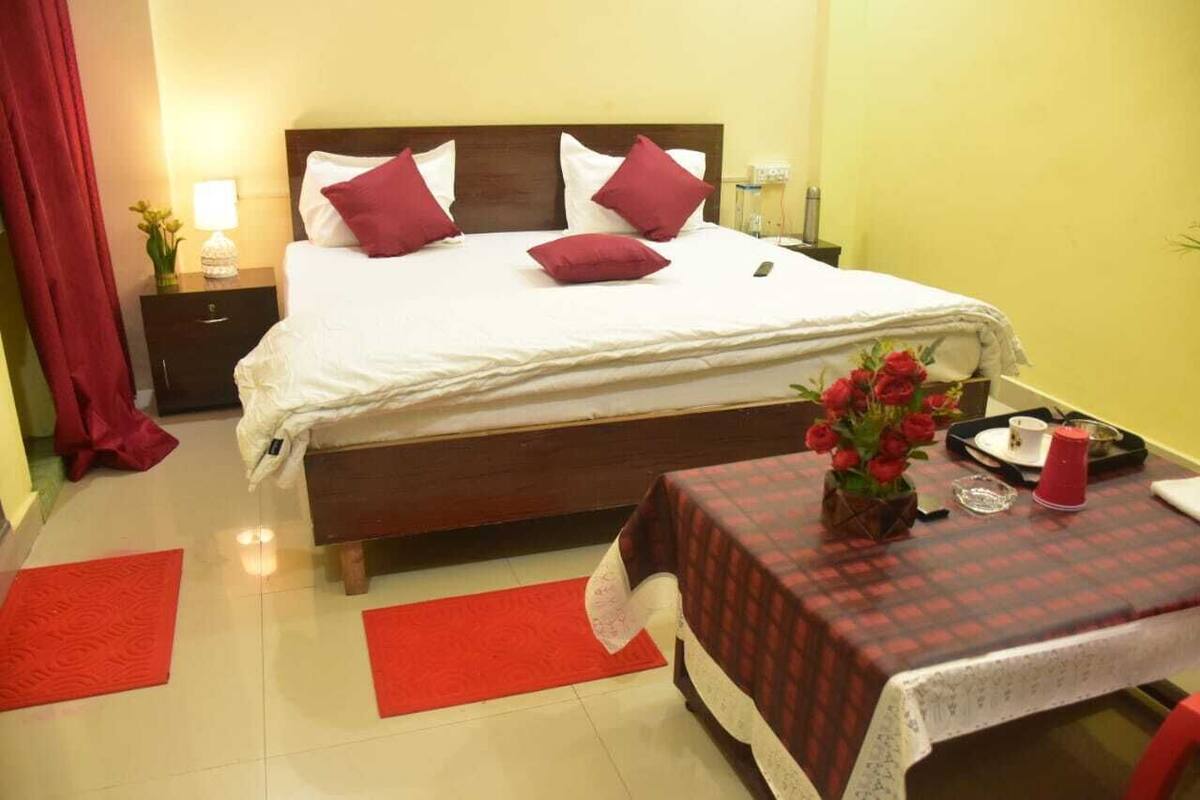 Una cama bien tendida con ropa de cama blanca y roja, una mesita de noche con una lámpara y una pequeña mesa con un jarrón de flores.