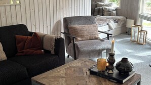 Ein gemütliches Wohnzimmer mit einem dunklen Sofa, einem hellen Sessel, einem Holztisch und einem Bett mit Blick durch ein Fenster.