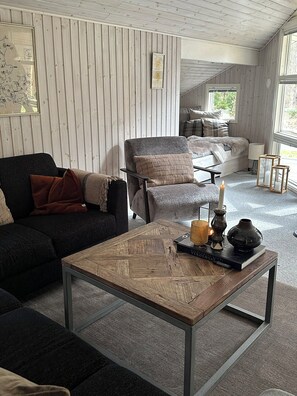 A cozy living room with a dark sofa, a light-colored armchair, a wooden coffee table, and a bed with a view through a window.