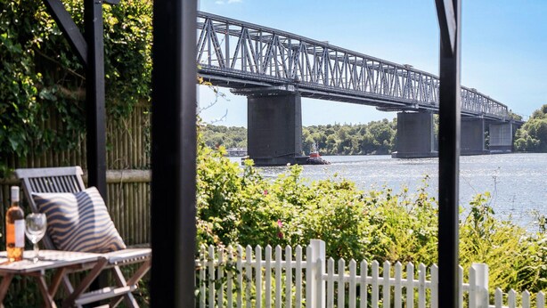 Blick auf eine Brücke über dem Wasser, einen weißen Zaun und Sitzgelegenheiten im Freien mit einer Flasche und einem Glas.