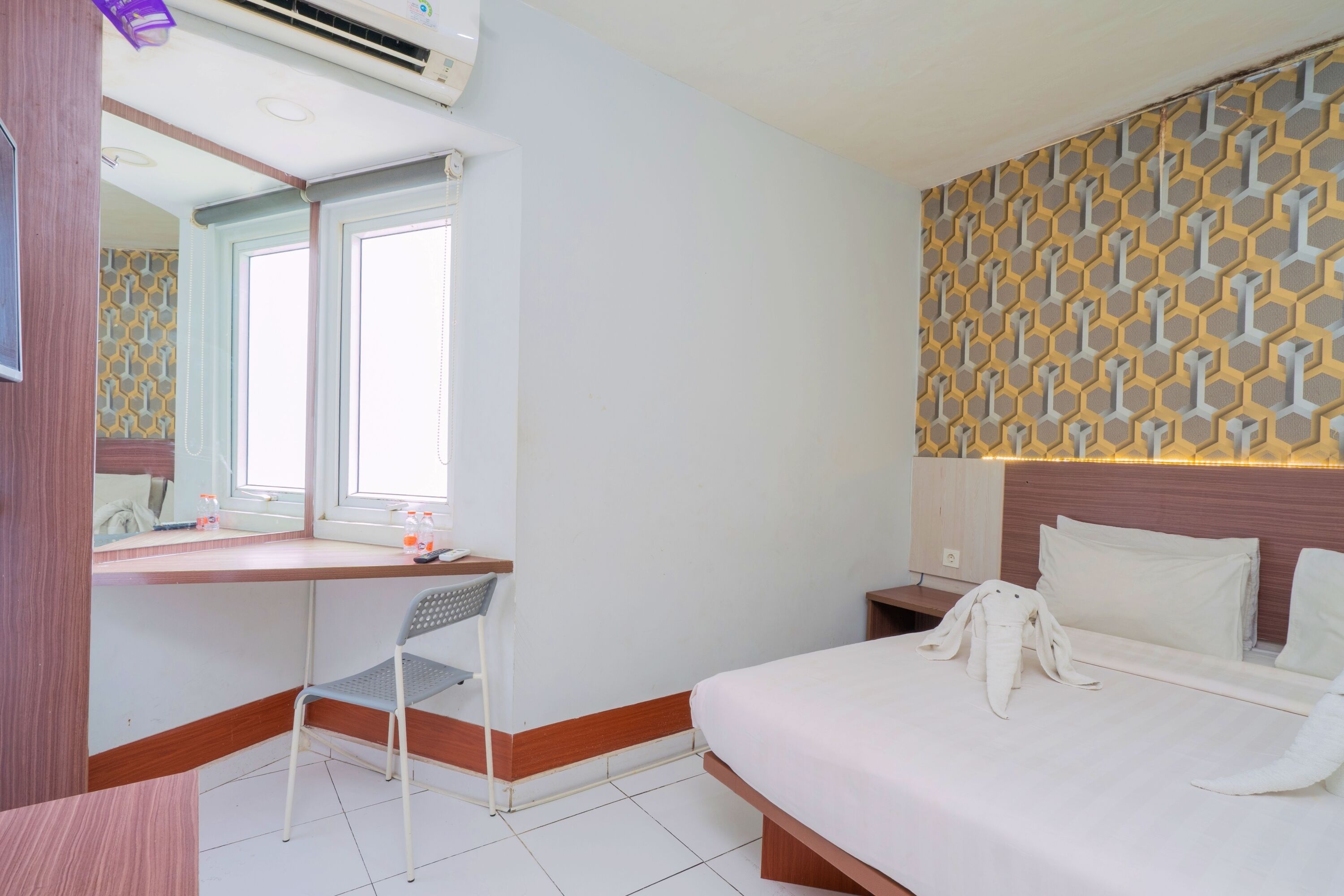 A hotel room with a bed, a desk with a chair, a window, and a patterned wall.
